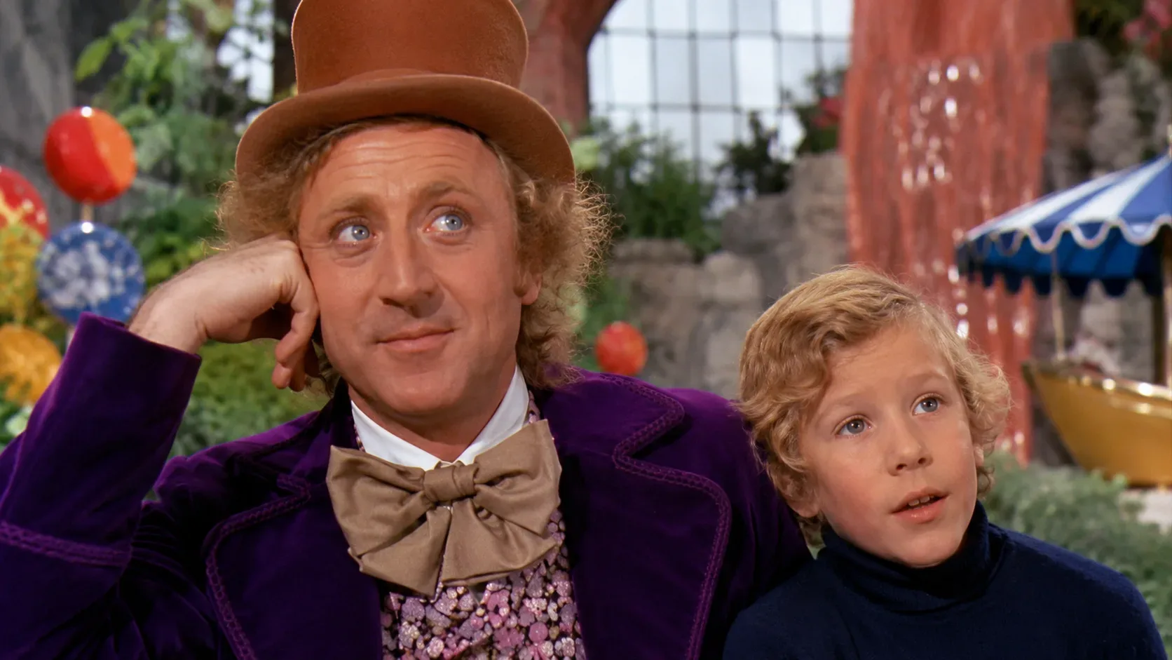 Willy Wonka de cartola marrom e casaco roxo ao lado de Charlie loiro, diante de um cenário colorido da fábrica de doces.
