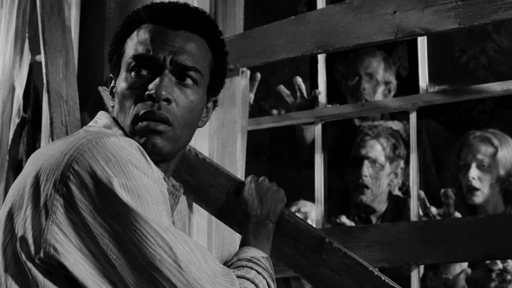 Cena de terror em preto e branco: homem negro segura uma tábua em uma casa escura diante de uma janela protegida por madeira, enquanto figuras cadavéricas tentam entrar.