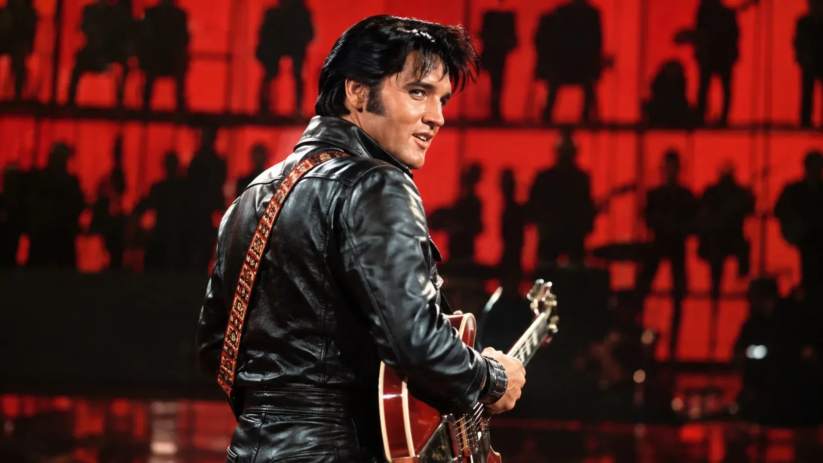 Elvis Presley em traje de couro preto com guitarra elétrica vermelha, diante de cenário iluminado em vermelho e silhuetas ao fundo.